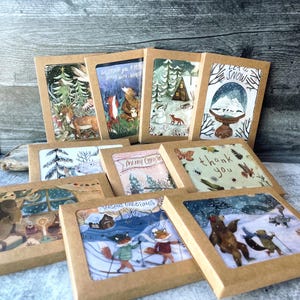 May include: A set of ten illustrated holiday greeting cards featuring winter scenes with animals. The cards are packaged in brown envelopes with a clear window to view the artwork. The cards feature a variety of winter scenes, including a snowy forest, a cabin in the woods, a snowman, and a group of animals skiing. The text on the cards includes "Wishing you a year filled with wonder", "Merry Christmas", "Thank you", and "Seasons Greetings".