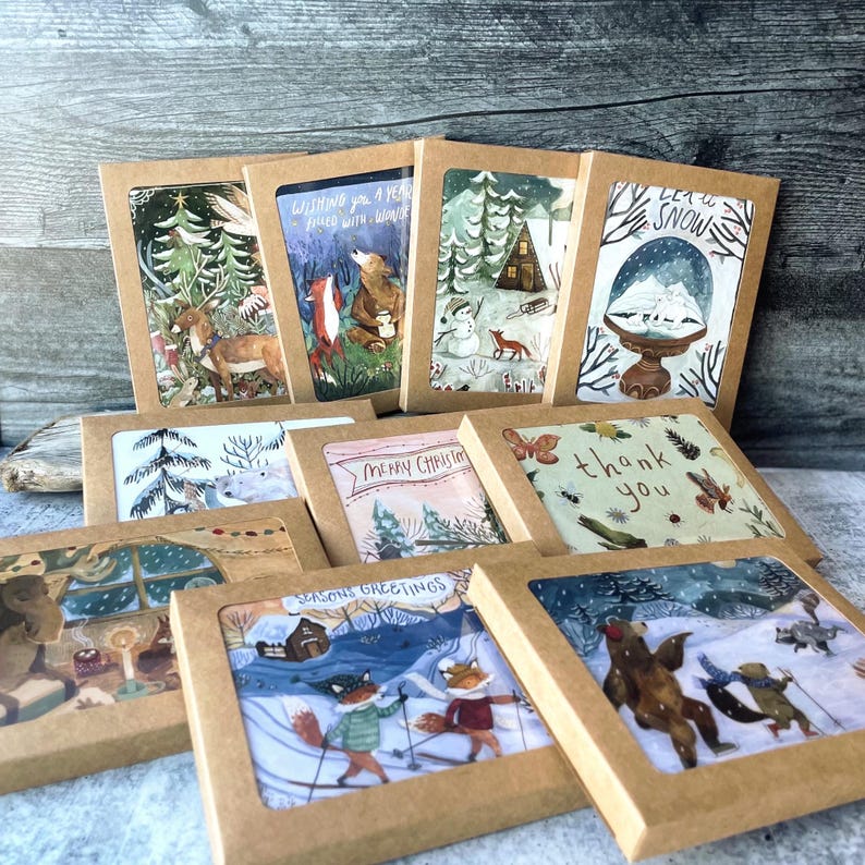 May include: A set of ten illustrated holiday greeting cards featuring winter scenes with animals. The cards are packaged in brown envelopes with a clear window to view the artwork. The cards feature a variety of winter scenes, including a snowy forest, a cabin in the woods, a snowman, and a group of animals skiing. The text on the cards includes "Wishing you a year filled with wonder", "Merry Christmas", "Thank you", and "Seasons Greetings".
