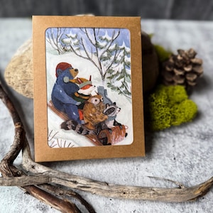 May include: A greeting card featuring a winter scene with a blue bear, fox, owl, and raccoon riding a sled. The illustration is set against a snowy backdrop with trees. The card is in a brown paperboard sleeve.
