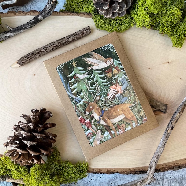 May include: A brown paper envelope with a Christmas card inside. The card features a colorful illustration of a deer, an owl, and a snowy forest scene.