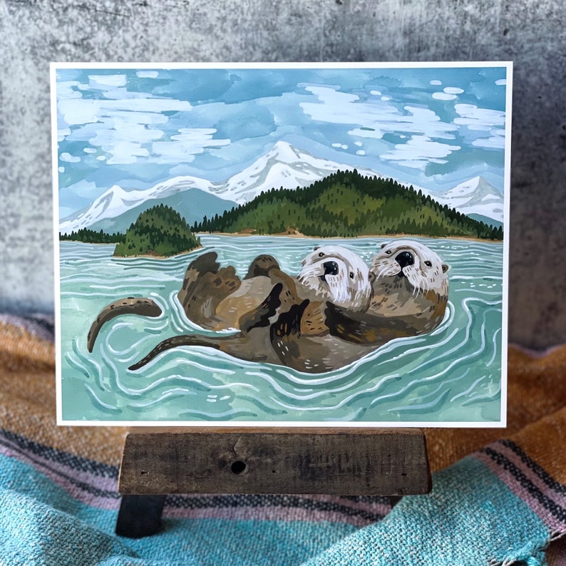 May include: Two sea otters, one brown and one gray, are floating in a turquoise blue body of water. The otters are cuddling and have their heads above the water. The background is a watercolor painting of a mountain range with snow-capped peaks and a green forest.