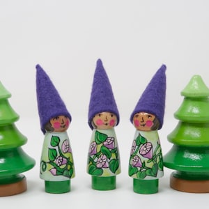 May include: Wooden gnome figurines with purple felt hats and painted faces stand between two green wooden Christmas trees. The gnomes wear white dresses with floral designs. The trees have brown bases.
