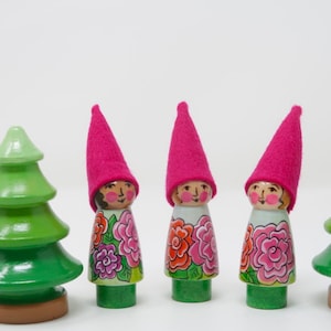 May include: Three wooden dolls with pink felt hats and floral designs stand in a row. Two green wooden trees are on either side of the dolls.