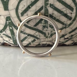 May include: A circular ring with a matte silver finish featuring five small, evenly spaced gold-toned spherical accents attached to the outer edge of the band. The ring has a smooth, consistent thickness throughout its entire circumference.