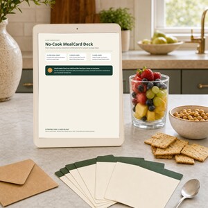 May include: A tablet displaying a No-Cook Meal Card Deck information page, accompanied by a set of cream-colored cards with dark green top borders, a brown paper envelope, and a silver spoon on a countertop next to fruit and crackers.