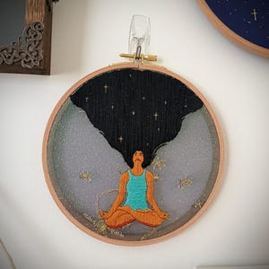 May include: Embroidered artwork in a wooden hoop. The design features a person in a seated yoga pose with long black hair that blends into a starry night sky. The person wears a turquoise top and orange pants. The background is gray.