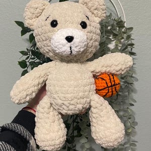 May include: A crocheted cream-colored teddy bear holding an orange basketball. The bear has black eyes and a white nose.