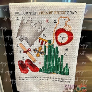 May include: A white kitchen towel with a graphic of the Wizard of Oz's Yellow Brick Road. The graphic includes a crown, a heart, a pair of ruby slippers, a diploma, and a green emerald city. The text "Follow the Yellow Brick Road" is at the top of the towel. The numbers 1 through 6 are next to each item, with the corresponding item's name listed below.