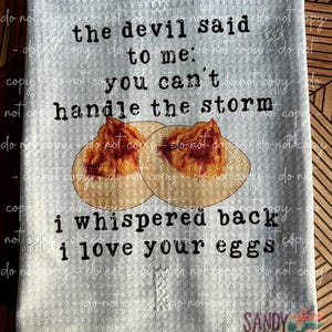 May include: A gray and white kitchen towel with the text "The devil said to me: You can't handle the storm. I whispered back I love your eggs."  Two fried eggs are pictured in the center of the towel.