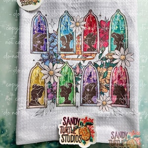 May include: A light gray towel featuring silhouettes of characters in glass bell jars, surrounded by flowers and a golden crown. The towel has the text "SANDY TURTLE STUDIOS" and a turtle logo.