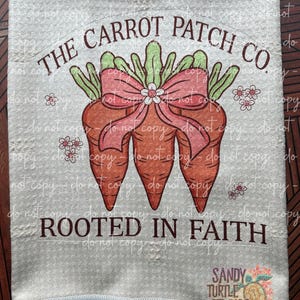May include: A white kitchen towel with a brown text overlay that reads "THE CARROT PATCH CO. ROOTED IN FAITH". The text is surrounded by a cartoon image of three carrots with green tops and a pink bow. The carrots are surrounded by small white flowers.
