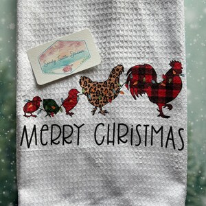 May include: White kitchen towel with a red and black plaid rooster, a leopard print hen, and three red cardinals holding Christmas lights. The text "Merry Christmas" is printed below the animals.