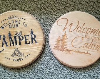 May include: Two wooden round signs with a rustic wood grain finish. The sign on the left says "Welcome to our Camper" with a small camper image and a campfire image. The sign on the right says "Welcome to our Cabin" with a stylized image of pine trees.