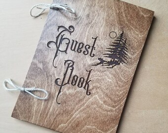 May include: A wooden guest book with a rustic brown finish. The cover is engraved with the words "Guest Book" in a decorative font and a stylized pine tree design.