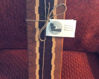 May include: A wooden cutting board with a brown, black, and light wood design. The board is tied with twine and has a tag that reads "The Rustic Woodworker" and "Handmade in Rochester Hills, Michigan, USA".