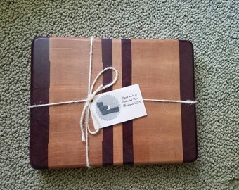 May include: A wooden cutting board with a natural wood finish and purple wood accents. The board is tied with twine and has a tag that reads "Hand made in Rochester Hills, Michigan USA".