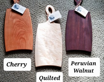 May include: Three wooden cutting boards with handles. The boards are made of cherry, quilted maple, and Peruvian walnut. The boards are all different sizes and shapes.