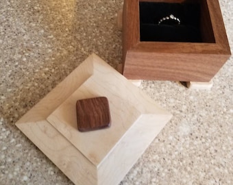 May include: A wooden ring box with a light wood lid and a dark wood base. The lid has a small square of dark wood in the center. The box is open and a silver ring is inside.