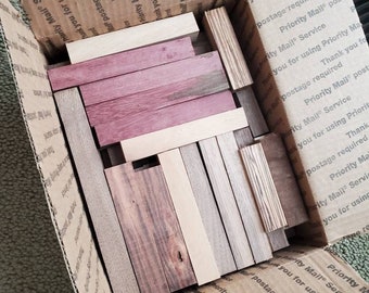 May include: A cardboard box filled with various wooden dowels in different colors and wood grains. The dowels are arranged in a random pattern and are all approximately the same length.
