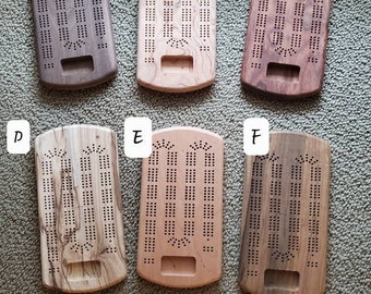 May include: Six wooden cribbage boards in various wood grains: Walnut, Gummy Cherry, Granadillo, Spalted Hackberry, Cherry, and Black Limba. Each board has a rectangular shape with drilled holes for pegs and a recessed area. The text "In Process" is also visible.