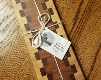 May include: A rectangular wooden cutting board with a checkered pattern of light and dark wood. The board is wrapped in twine and has a tag that reads "Hand made in Rochester Hills, Michigan, USA".