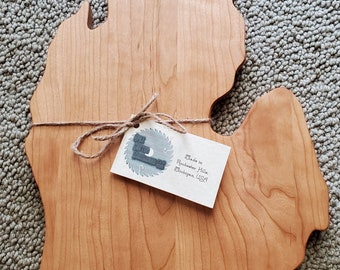 May include: A wooden cutting board shaped like the state of Michigan. The board is made of cherry wood and has a natural finish. It is tied with twine and has a tag that reads "Made in Rochester Hills, Michigan, USA".