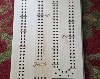 May include: A wooden game board with a track for playing a game of cribbage. The board has a light wood finish and features the words "Start" and "Finish" along with numbers 30, 60, and 90. The board has a small compartment for storing game pieces.