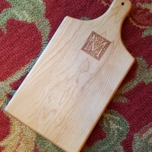 May include: A light wood cutting board with a handle. The board has a decorative engraved letter "M" in the center.