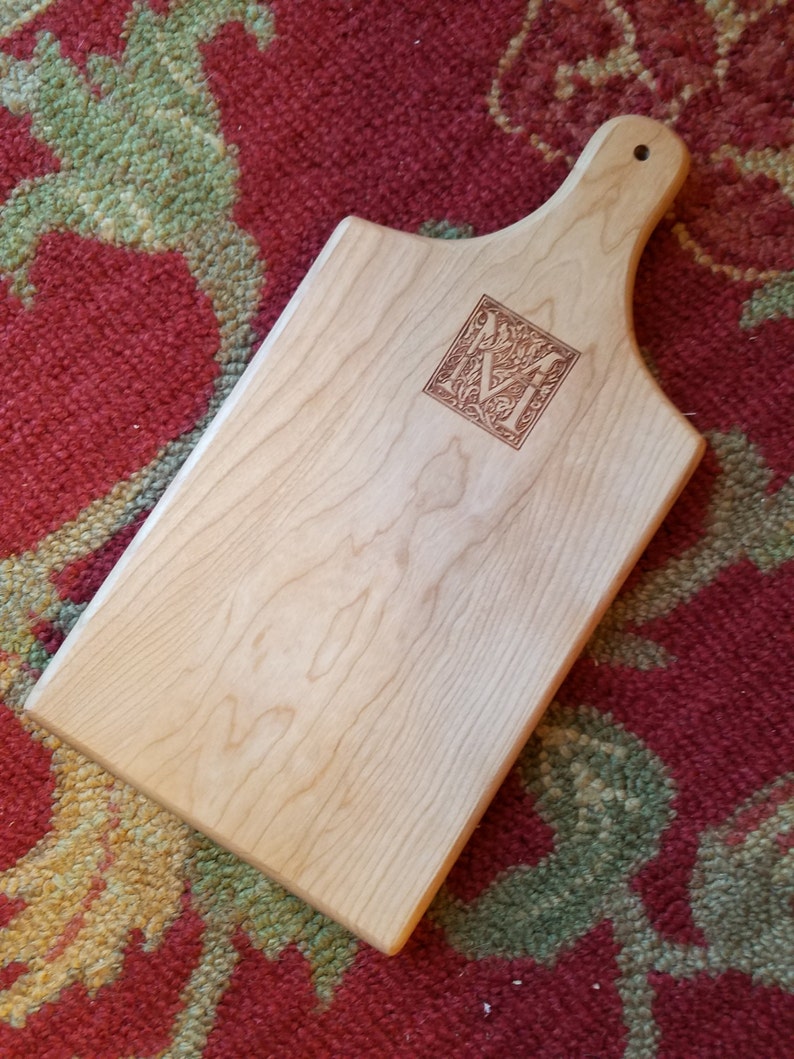 May include: A light wood cutting board with a handle. The board has a decorative engraved letter "M" in the center.