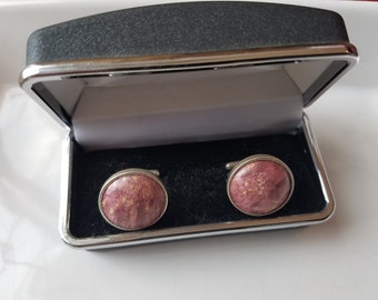 May include: A pair of round cufflinks in a black velvet box. The cufflinks are silver with a pink and gold marbled design.