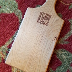 May include: A light wood cutting board with a handle. The board has a decorative letter "D" burned into the surface.