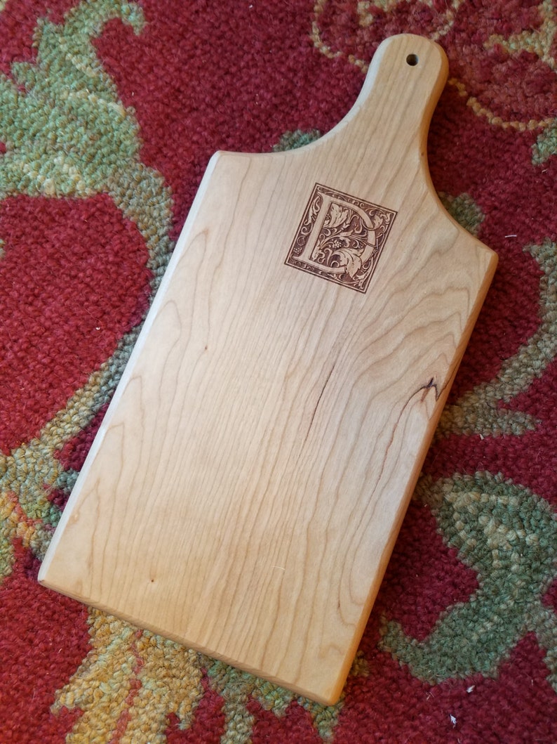 May include: A light wood cutting board with a handle. The board has a decorative letter "D" burned into the surface.