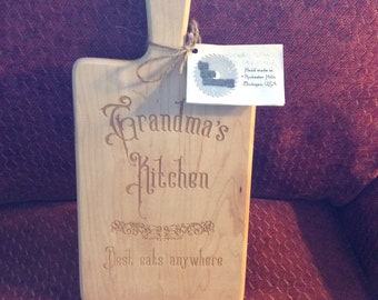 May include: A wooden cutting board with a handle, engraved with the text "Grandma's Kitchen Best eats anywhere". The board has a small tag attached with the text "Hand made in Rochester Hills, Michigan, USA".