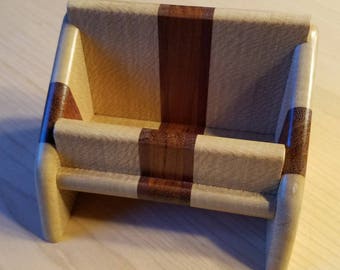 May include: A small wooden box with a bench-like design. The box is made of light and dark brown wood, with a rectangular shape and rounded edges. The front and back have a dark brown stripe. The box is likely used for holding small items.