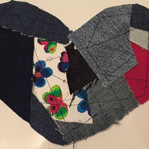 Könnte beinhalten: Eine herzförmige Patchwork-Decke aus Jeansstoff in verschiedenen Blautönen, Schwarz und Grau. Die Mitte der Decke zeigt einen weißen Stoff mit einem bunten Schmetterlingsmuster.