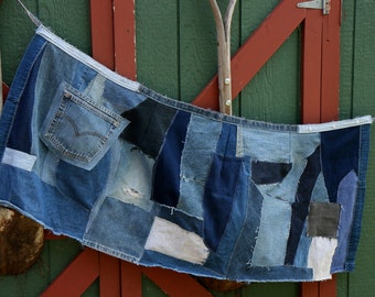May include: A denim apron made from repurposed blue jeans. The apron has a large pocket on the front and is held up by a white strap.