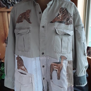 May include: A beige button-up shirt with a patchwork design. The shirt features various animal appliques, including a leopard, tiger, lion, and llama. The shirt has a large pocket on the left side and a smaller pocket on the right side.