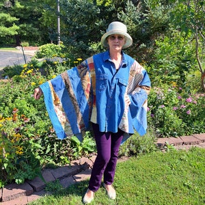 May include: A woman wearing a blue denim shirt with a patchwork design, a white hat, purple pants, and white shoes. She is standing in a garden with green grass and flowers.