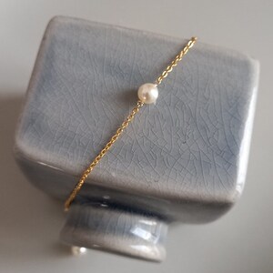 Puede incluir: Una delicada pulsera de cadena dorada con una sola perla blanca y redonda. La pulsera est&aacute; colocada sobre un objeto de cer&aacute;mica azul claro texturizado. El fondo es de color neutro.