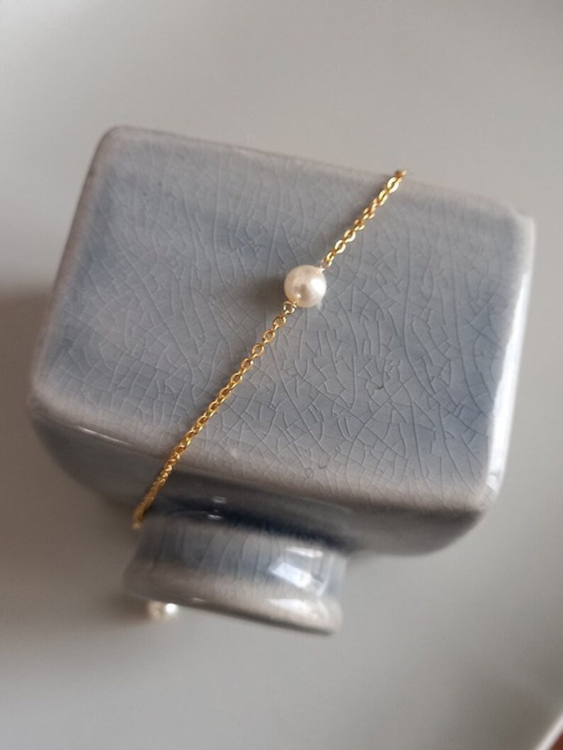 Puede incluir: Una delicada pulsera de cadena dorada con una sola perla blanca y redonda. La pulsera est&aacute; colocada sobre un objeto de cer&aacute;mica azul claro texturizado. El fondo es de color neutro.