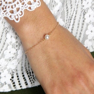 Puede incluir: Una delicada pulsera de cadena dorada con una sola perla redonda. La pulsera se lleva en la mu&ntilde;eca, con una manga de encaje blanco visible en el fondo. La pulsera es una pieza de joyer&iacute;a sencilla y elegante.