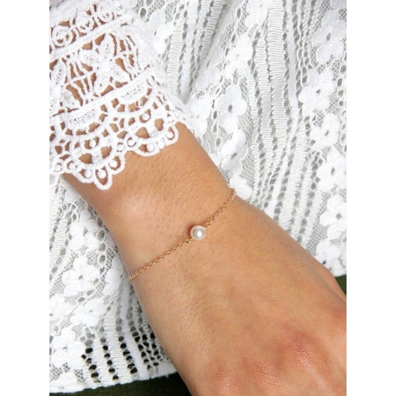 Puede incluir: Un delicado brazalete de cadena dorada con una sola perla redonda. El brazalete es simple y elegante, con una fina cadena y una peque&ntilde;a perla brillante como punto focal. El brazalete se muestra en una mu&ntilde;eca.