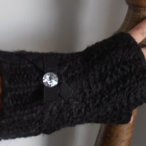 May include: A black knitted fingerless glove with a small black bow and a clear, round jewel. The glove is made of a thick, textured yarn. The bow is centered on the glove, and the jewel is in the center of the bow.