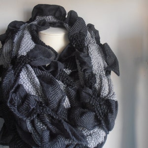 以下が含まれることがあります： グレーとブラックのニットスカーフで、フリルデザインになっています。スカーフは柔らかく、質感のある糸で作られています。