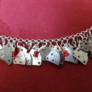 May include: A silver charm bracelet with multiple watch part charms, each with different text, including "ELGIN WATCH CO", "SEVENTEEN 17 JEWELS", "TIME", and "JATA JEWELS". Each charm has a small red bead attached.
