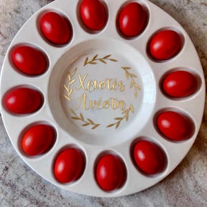 May include: A white ceramic plate with 10 red Easter eggs arranged around the edge. The center of the plate has gold lettering and a gold leaf design. The text reads "Christos Anesti".