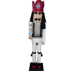 May include: A wooden nutcracker figurine in a regal design. It features a red crown with blue and clear gems, a black mustache, and long black hair. The uniform is white with blue and gold accents. The figurine is standing on a black base.