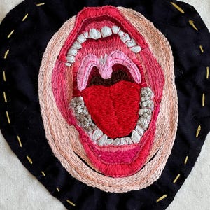 May include: A close-up of a black fabric circle with a detailed embroidered image of an open mouth with teeth and a tongue. The embroidery is done in shades of pink, red, and white.