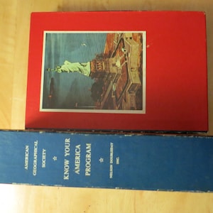 May include: A red box with an image of the Statue of Liberty and a blue box with the text "KNOW YOUR AMERICA PROGRAM" are on a wooden surface. The boxes appear to be vintage.