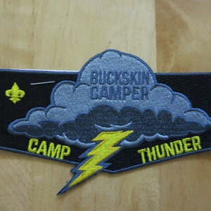 May include: Embroidered patch with a black background, featuring a cloud with the text "BUCKSKIN CAMPER" and a yellow lightning bolt. The patch also includes the words "CAMP THUNDER" and a gold fleur-de-lis.